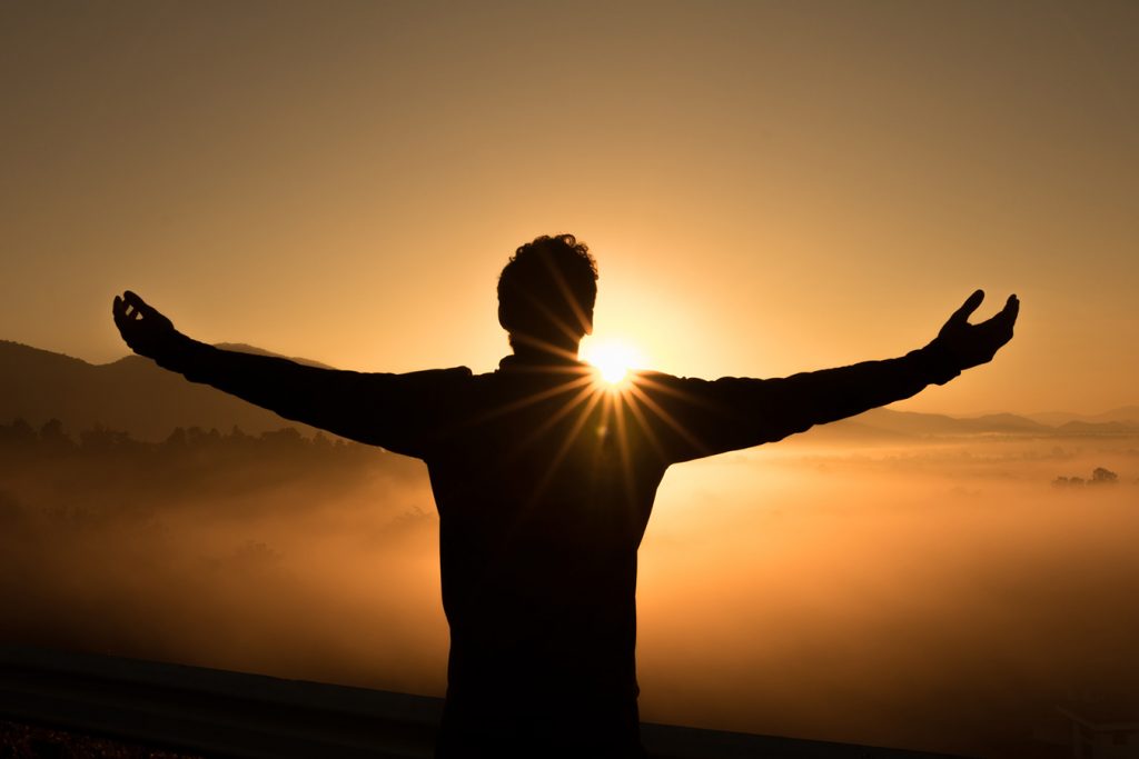 Man standing at sunset with arms outstretched offering hope with the orange yellow sun setting in front of him with foggy clouds in the distance.