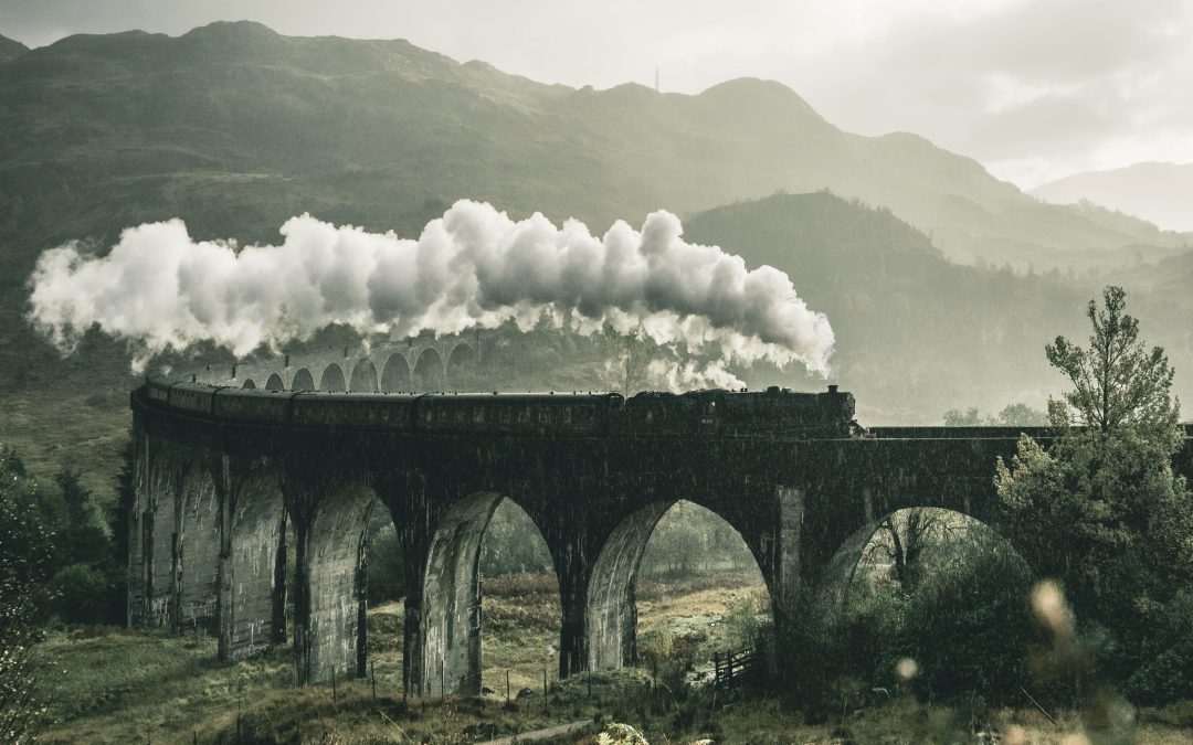 old black and white sepia train on bridge train tracks with clouds of billowing smoke in the sky and mountains in the distance