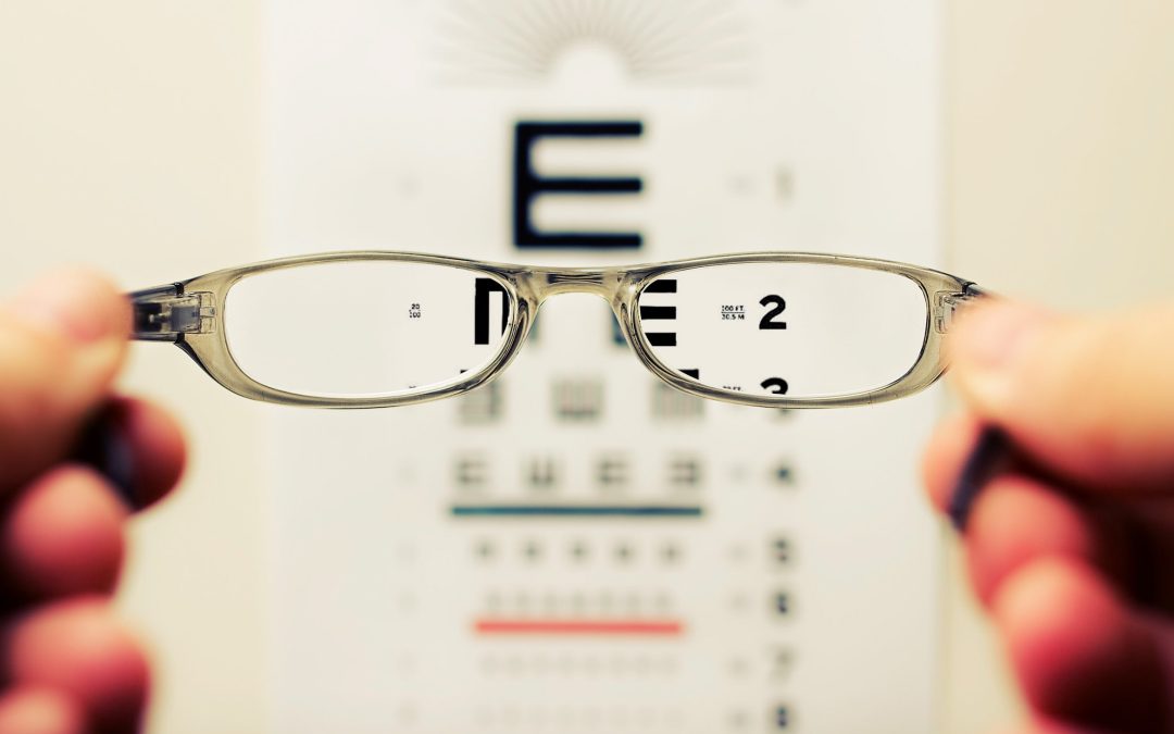 Part of a hand holding a pair of reading glasses and facing an eye chart with letters some blurry and others in focus