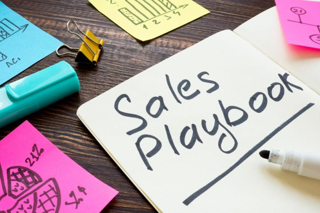 book laying on an office work table that says sales playbook next to post-its and other business ideas and strategies which lead to team work 